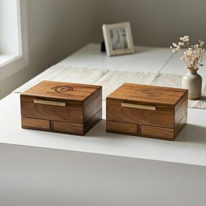 May include: Two rectangular wooden boxes with a dark brown grain, each featuring a horizontal gold metal handle on the lid. The left box is engraved with a crest and the text Thomas Anderson, while the right box displays a circular floral design.