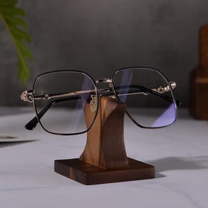 Puede incluir: Un par de gafas cuadradas negras con detalles dorados en un soporte de madera. Las gafas tienen lentes tintadas de azul.