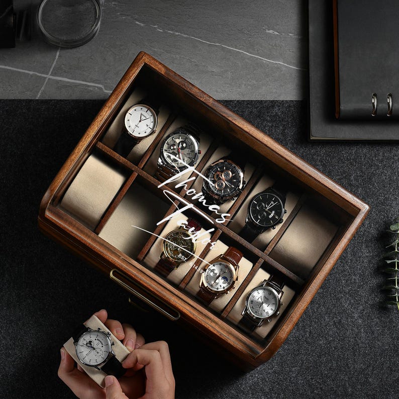 Op de afbeelding: Een houten horlogedoos met acht horloges. De horloges hebben verschillende band- en wijzerplaatontwerpen. De doos heeft een glazen bovenkant en een messingkleurige handgreep. De tekst "Thomas Taylor" staat op de doos.