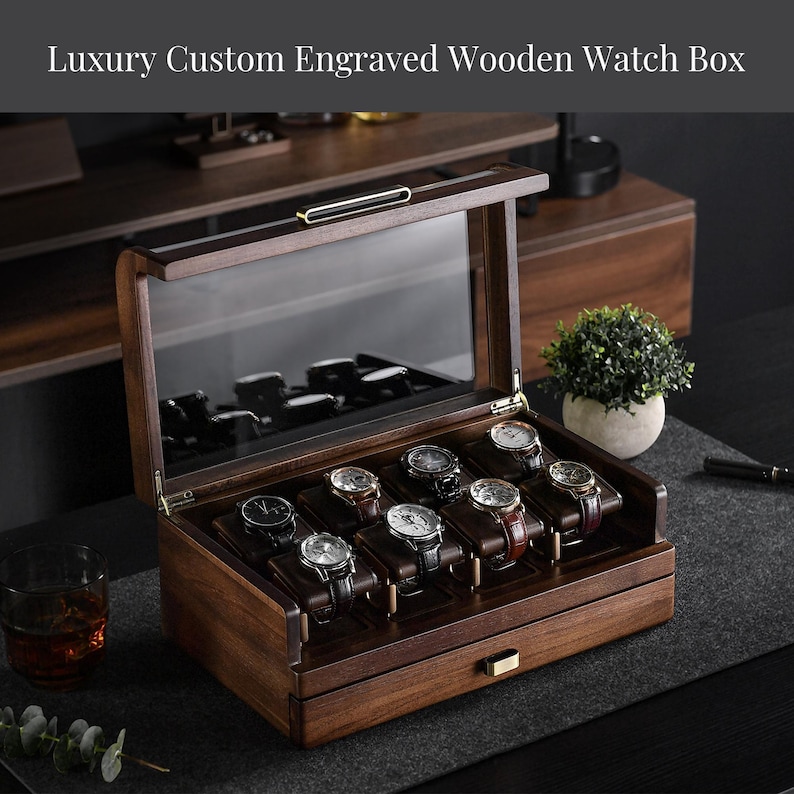 A wooden watch box with multiple compartments, displaying a variety of watches, placed on a wooden surface with a potted plant and a glass of what appears to be a beverage nearby.