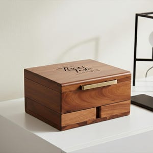 Puede incluir: Una caja de joyer&iacute;a de madera con forma rectangular y dos cajones. La caja es de color marr&oacute;n c&aacute;lido con un asa dorada. La parte superior de la caja tiene el nombre "Thomas Taylor" grabado en cursiva.