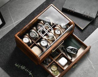 May include: A wooden watch storage box with a glass lid featuring a white logo, containing six wristwatches on beige cushions. A pull-out drawer below holds a car key fob, a white wireless earbud case, and two gold-toned circular coins.