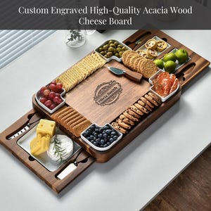 May include: Custom engraved acacia wood cheese board with a variety of snacks. The board features a central cutting surface, surrounded by small white dishes filled with olives, grapes, and cookies. The text "Olivia Smith" is engraved on the board.