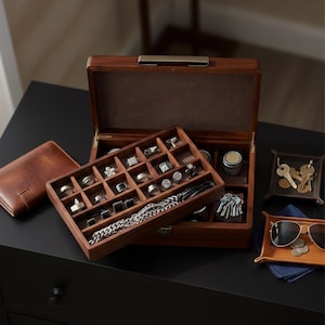 Puede incluir: Una caja de joyería de madera oscura abierta, que revela anillos, gemelos y una cadena. También se ven una cartera de cuero y pequeñas bandejas con llaves, monedas y gafas de sol.