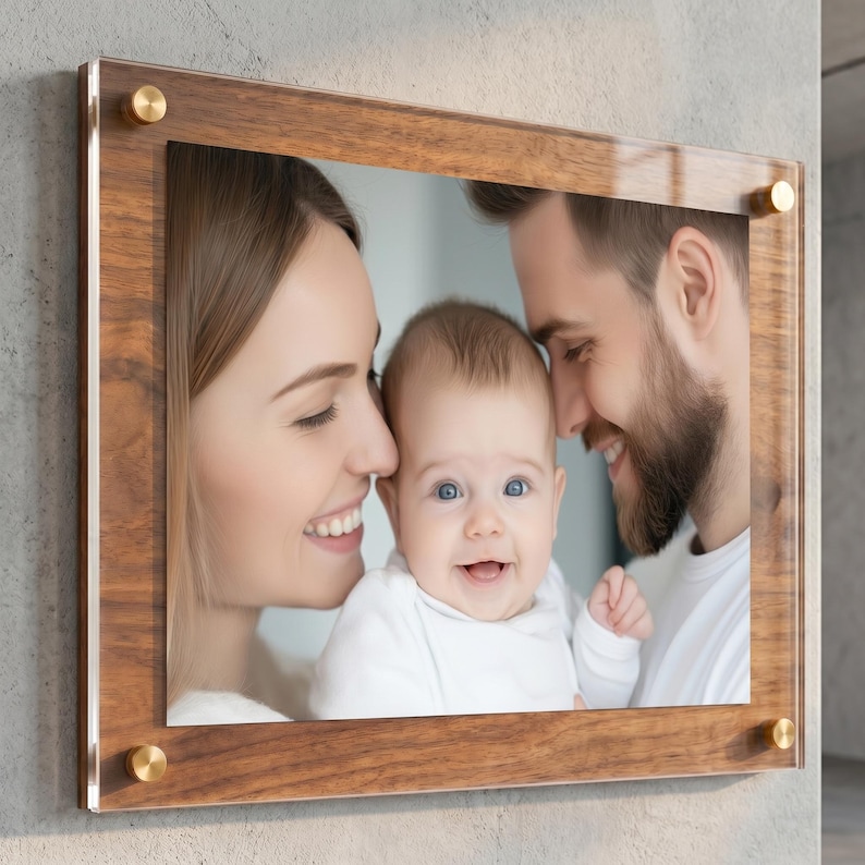 May include: A rectangular photo frame featuring a dark wood border and a clear acrylic front panel. The frame is secured at each corner with circular gold-toned metal hardware. Inside the frame is a photograph of two adults and a baby.