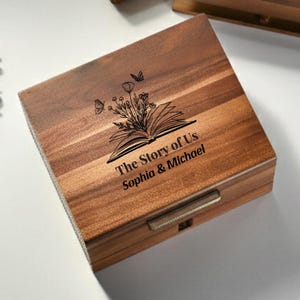 Puede incluir: Caja de madera con tapa con bisagras, con un diseño grabado con láser de un libro abierto, flores y mariposas. El texto "The Story of Us" y los nombres "Sophia & Michael" están debajo. Acabado de veta de madera natural.