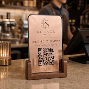 May include: A rectangular rose gold sign with a black frame held in a dark wood base. The sign features the text SOLACE COFFEE, ENJOYED YOUR VISIT?, SCAN TO LEAVE A REVIEW, a large QR code, and decorative floral line art elements.