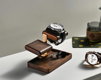 May include: A dark wood watch stand with a gold-toned metal support, featuring a padded top bar for a wristwatch, a flat platform for accessories, and a pair of black square cufflinks resting on the base, alongside a separate wristwatch.