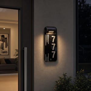 May include: A rectangular, black, wall-mounted house number sign featuring the illuminated digits 777 in a vertical arrangement. The sign includes a built-in solar light at the top that casts a warm glow downwards, highlighting the silver-coloured numbers.