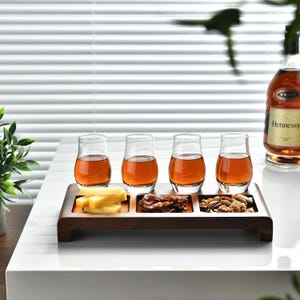 A wooden tray with four glasses of amber-colored liquid, likely whiskey, and various snacks such as cheese, nuts, and crackers, accompanied by a bottle of Hennessy on a white table.