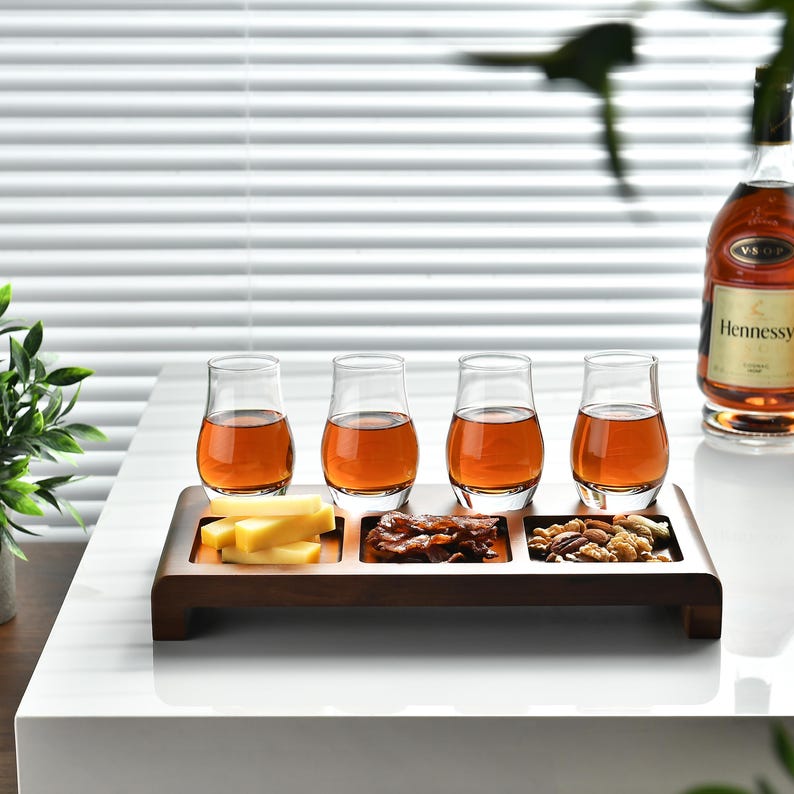 A wooden tray with four glasses of amber-colored liquid, likely whiskey, and various snacks such as cheese, nuts, and crackers, accompanied by a bottle of Hennessy on a white table.