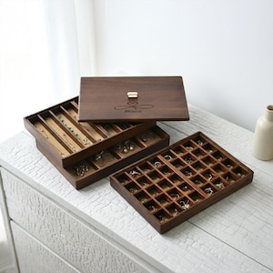 Puede incluir: Una caja de joyería de madera marrón oscuro con múltiples compartimentos y una tapa con bisagras. La caja tiene un cierre dorado y está grabada con el nombre "Maria" y un símbolo de corazón. La caja está abierta, revelando collares, anillos y pendientes.