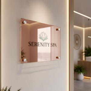 Puede incluir: Un letrero rectangular de color oro rosa para "Serenity Spa" está montado en una pared de color crema. El letrero presenta el nombre del spa en una fuente serif, junto con un logotipo de flor de loto. El letrero se sujeta con cuatro fijaciones circulares plateadas.