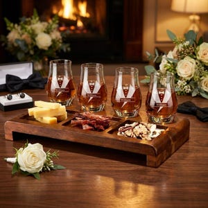 A wooden tray with four glasses of whiskey, a plate of cheese, and some nuts, all set against a backdrop of a fireplace and flowers.