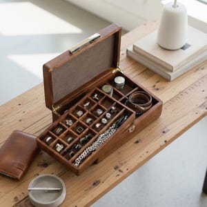 May include: A rectangular wooden jewelry box with a hinged lid and a multi-compartment interior. The box features a dark brown finish, a metal latch, and a felt-lined interior containing various rings, a silver chain, and stacked coins.