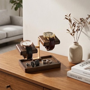 May include: A wooden watch display stand with two watches, a black leather strap watch and a brown leather strap watch. The stand also holds a pair of cufflinks and two rings. The stand is on a wooden surface.