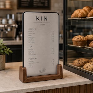 May include: A vertical rectangular menu sign with a black frame and a wooden base. The sign displays the text KIN COFFEE followed by lists of coffee, tea, and pastry items with prices. A small line drawing of a leafy branch is at the bottom.