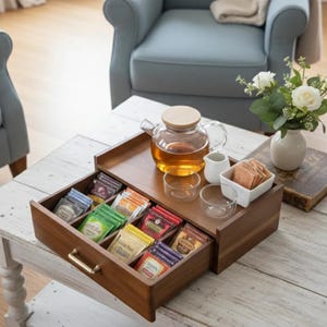 May include: A wooden tea storage box with a pull-out drawer containing various tea bags in colorful wrappers. On top sits a glass teapot with a wooden lid, two small glass cups, a white ceramic creamer, and a white bowl filled with tea bags.