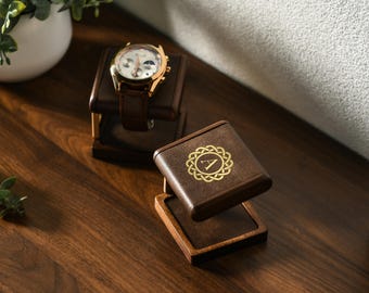 May include: Two dark brown wooden watch stands with a square base and a raised platform. One stand holds a gold-tone wristwatch with a brown leather strap. The other stand features a gold circular emblem with the letter A in the center.