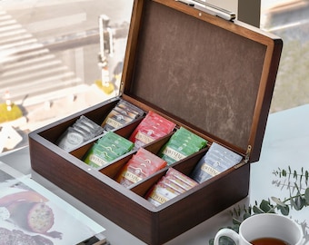 May include: A dark wooden tea box with a hinged lid, filled with various tea bags. The tea bags are in different colors, including green, red, and silver. A white mug of tea sits nearby. The box is open, revealing the tea bags.