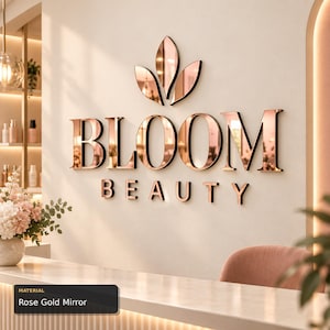 Könnte beinhalten: Ein Wandschild mit dem Schriftzug BLOOM BEAUTY in reflektierenden roségoldenen Spiegelbuchstaben. Über dem Text befindet sich ein dreiblättriges Blumensymbol in derselben roségoldenen Ausführung. Die Buchstaben und das Symbol sind an einer hellen Wand.