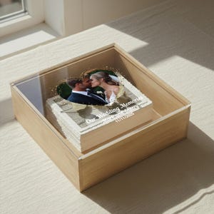 May include: A wooden box with a clear acrylic lid contains a stack of photos. The top photo features a couple kissing inside a heart-shaped frame. The text "Our Wedding Memories Mark and Sherry 11/11/2025" is printed on the photo.