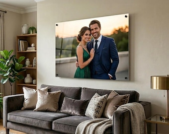 May include: A large, rectangular acrylic print of a couple in formal attire, mounted on a beige wall. The couple is embracing, with the woman in a green dress and the man in a navy suit. The print hangs above a gray sofa with decorative pillows.