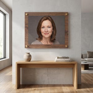 May include: A rectangular wooden picture frame with four gold-toned corner fasteners, displaying a portrait of a person with shoulder-length brown hair. The frame is mounted on a light gray wall above a light-colored wooden console table.