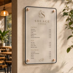 Op de afbeelding: Een rechthoekig menubord met een zwarte rand en vier koperkleurige afstandhouders. Het bord bevat de tekst Solace Coffee, een lijst met koffie, thee en gebak met prijzen, en florale illustraties op een lichtbeige ondergrond.