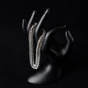 May include: A silver chainmail bracelet displayed on a black hand-shaped jewelry holder. The bracelet is made of small, interlocking metal rings, creating a textured, flexible design. The background is a solid black, highlighting the bracelet's metallic sheen.