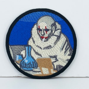 May include: Embroidered patch featuring a stylized figure with a white fur-like coat, red accents, and a cigarette. The patch has a blue background and a black border. A blue flask and a small table are also visible.