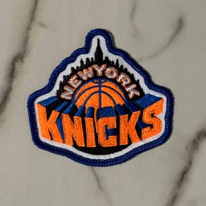May include: An embroidered patch featuring the New York Knicks logo with a basketball graphic, a black city skyline silhouette, and the text NEW YORK KNICKS in bold orange letters with blue outlines, all bordered by a thick blue and white stitched edge.