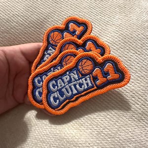 May include: A stack of three embroidered patches featuring a basketball graphic and the text CAPN CLUTCH 11. The patches have a black background, white lettering, orange borders, and orange numbers. The design includes a basketball icon.