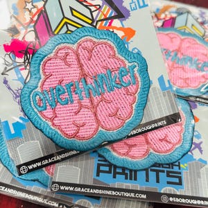 May include: A pink and blue embroidered brain patch with the text "overthinker" on it. The patch is on a white background with a city skyline design. The text "www.graceandshineboutique.com" and "@5boroughprints" are also visible.