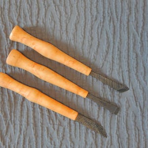 May include: Three wooden handled carving tools with black metal blades. The tools are laid out on a textured gray surface.