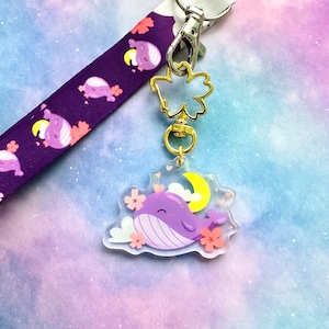 May include: Purple whale charm with a crescent moon and pink cherry blossoms on a clear background. The charm is attached to a gold chain and a purple lanyard with a white background and a repeating pattern of whales and cherry blossoms.