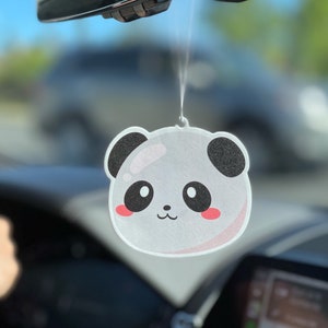 May include: A white panda bear car air freshener hanging from the rearview mirror of a car. The panda has black ears and eyes, a pink nose, and a pink blush on its cheeks.