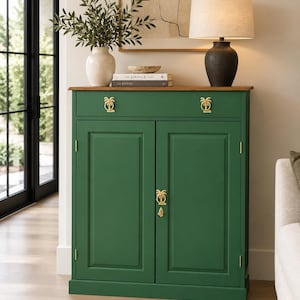 May include: A forest green wooden cabinet featuring two doors and one drawer with gold-toned palm tree-shaped hardware. The cabinet has a flat brown top surface and is accented with gold hinges on the doors, providing a storage solution.