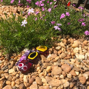 May include: A glass bee figurine with red, yellow, and black accents. The bee is sitting on a bed of small brown rocks and surrounded by purple flowers.