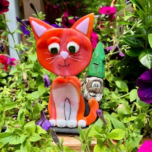 May include: A fused glass cat figurine with bright orange fur, white chest and paws, pink ears, and green eyes, positioned next to a small, carved wooden gnome figure with a green hat and a white beard, both standing upright.