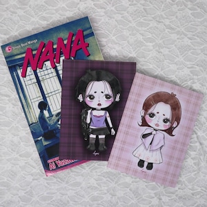 May include: A Nana Shojo Beat manga volume by Ai Yazawa alongside two rectangular art prints featuring stylized chibi-style character illustrations. One print shows a character in a purple top on a dark plaid background, the other in a light dress.