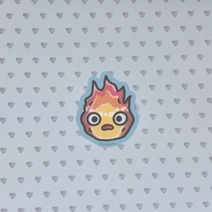 Puede incluir: Una pegatina de un esp&iacute;ritu de fuego de dibujos animados con una expresi&oacute;n de sorpresa. El esp&iacute;ritu es amarillo con llamas naranjas y rojas. La pegatina est&aacute; sobre un fondo azul claro con un patr&oacute;n de peque&ntilde;os corazones grises.