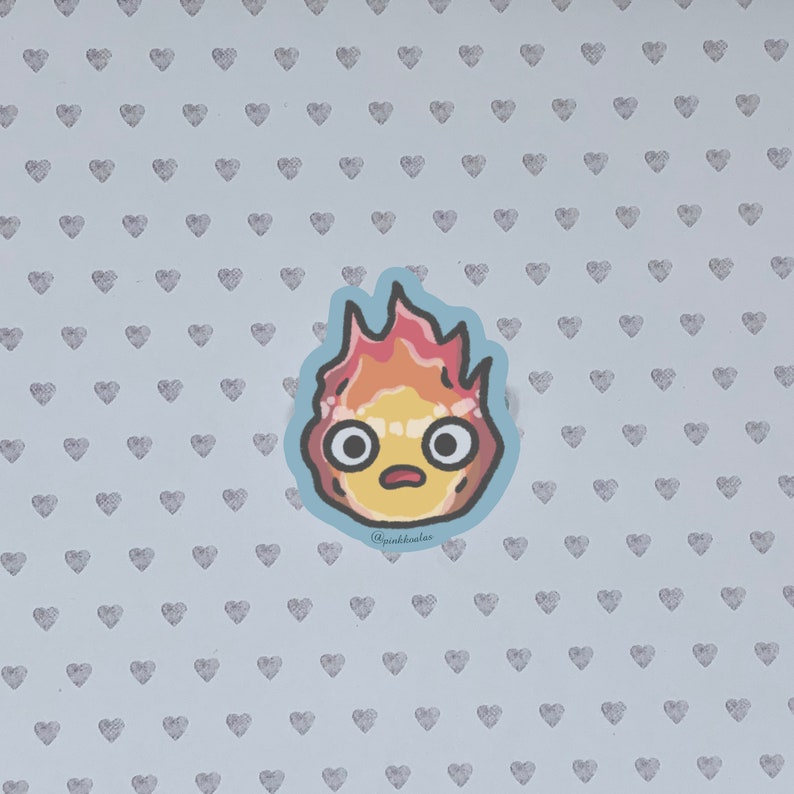 Puede incluir: Una pegatina de un esp&iacute;ritu de fuego de dibujos animados con una expresi&oacute;n de sorpresa. El esp&iacute;ritu es amarillo con llamas naranjas y rojas. La pegatina est&aacute; sobre un fondo azul claro con un patr&oacute;n de peque&ntilde;os corazones grises.