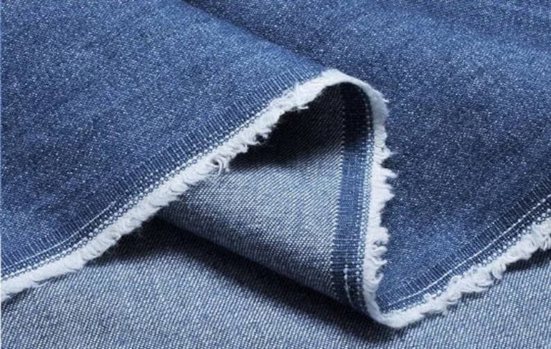May include: Close-up of a blue denim fabric with a raw edge. The fabric is folded over itself, showing the texture and weave of the material.