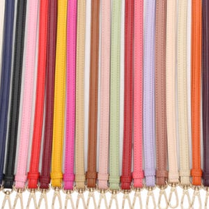 Könnte beinhalten: Eine Sammlung farbenfroher, schmaler Lederriemen mit goldfarbenen Metallverschlüssen. Die Riemen sind in verschiedenen Farben erhältlich, darunter Schwarz, Rosa, Rot, Gelb und Braun. Die Riemen sind wahrscheinlich für Handtaschen oder anderes Zubehör.