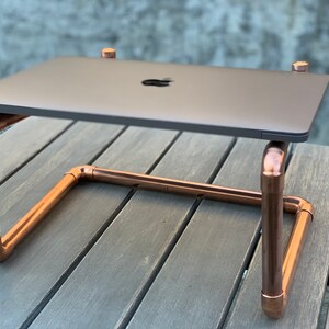 May include: A laptop stand constructed from polished copper pipes and fittings. The stand features a U-shaped base with four vertical supports that hold a dark gray laptop with a visible Apple logo on the lid, creating a raised platform.