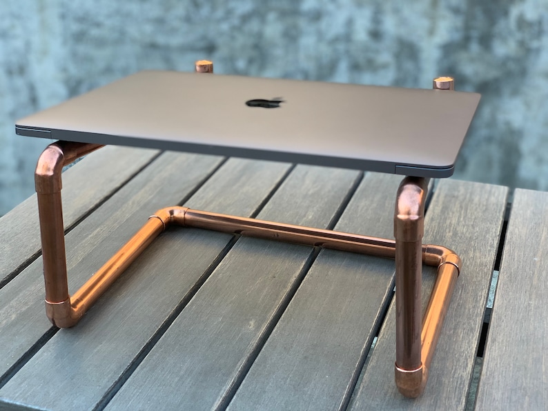 May include: A laptop stand constructed from polished copper pipes and fittings. The stand features a U-shaped base with four vertical supports that hold a dark gray laptop with a visible Apple logo on the lid, creating a raised platform.
