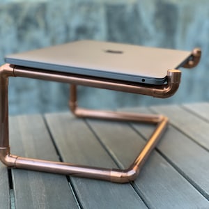 May include: A laptop stand constructed from polished copper pipes and fittings. The frame features a rectangular base with an elevated platform designed to hold a closed grey laptop. The metallic structure has a warm, reflective finish.