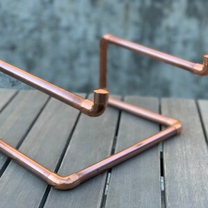 May include: A laptop stand constructed from polished copper piping. The frame features a rectangular base with two angled support arms extending upward, each ending in a small vertical hook to secure a device in place. The metallic finish is shiny.