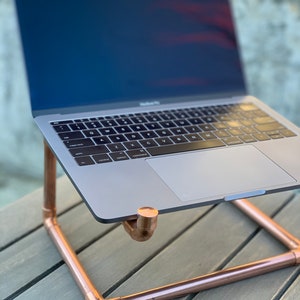 May include: A space gray laptop computer resting on a copper pipe stand. The stand is constructed from metallic copper-colored pipes and fittings, featuring a U-shaped base and two upward-facing supports that hold the laptop at an elevated angle.